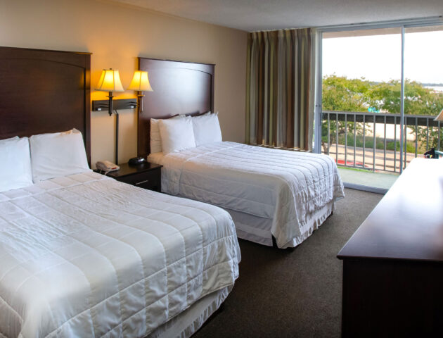 A spacious Yorktown hotel room with two full-sized beds, white linens, and a glass sliding door to a private balcony in Yorktown.