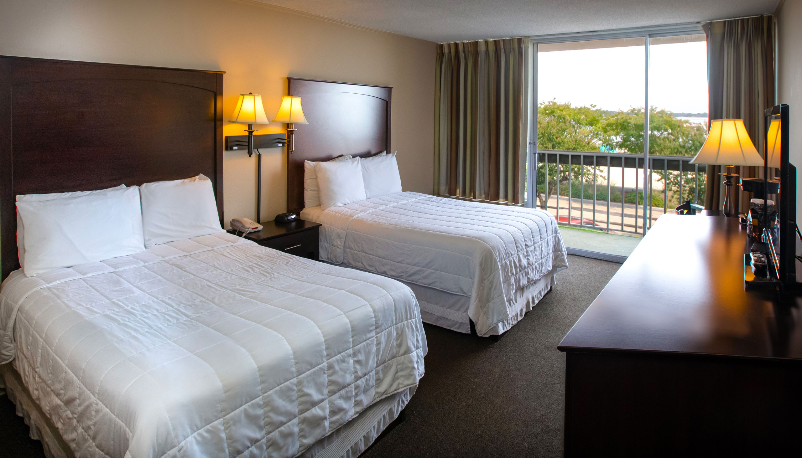A spacious Yorktown hotel room with two full-sized beds, white linens, and a glass sliding door to a private balcony in Yorktown.