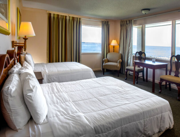 A spacious Yorktown hotel room with colonial green carpet, two queen beds with white linens, a desk, and expansive waterfront views.