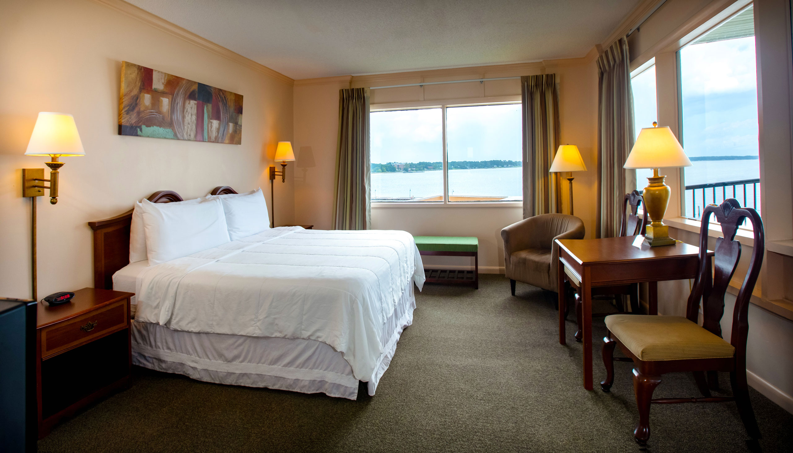 A spacious Yorktown hotel room with colonial green carpet, a king bed with white linens, a desk, and waterfront views.