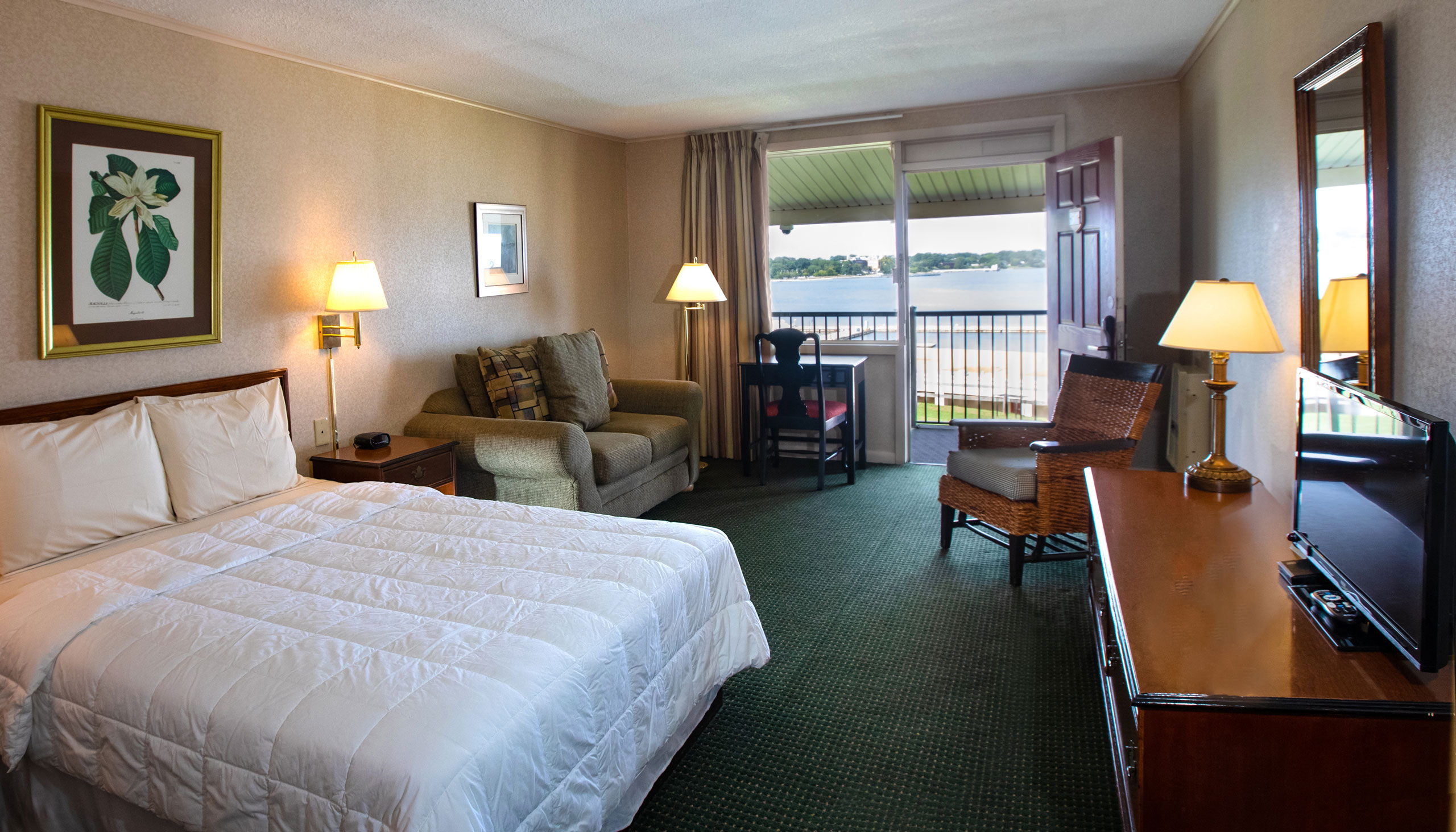 A spacious Yorktown hotel room with colonial green carpet, a queen bed, a love seat, desk, and terrace.