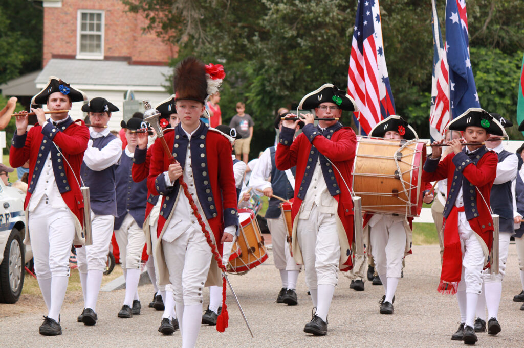 Revolutionary War reenactors with red coats, and a pipe & drum corps marching in a parade